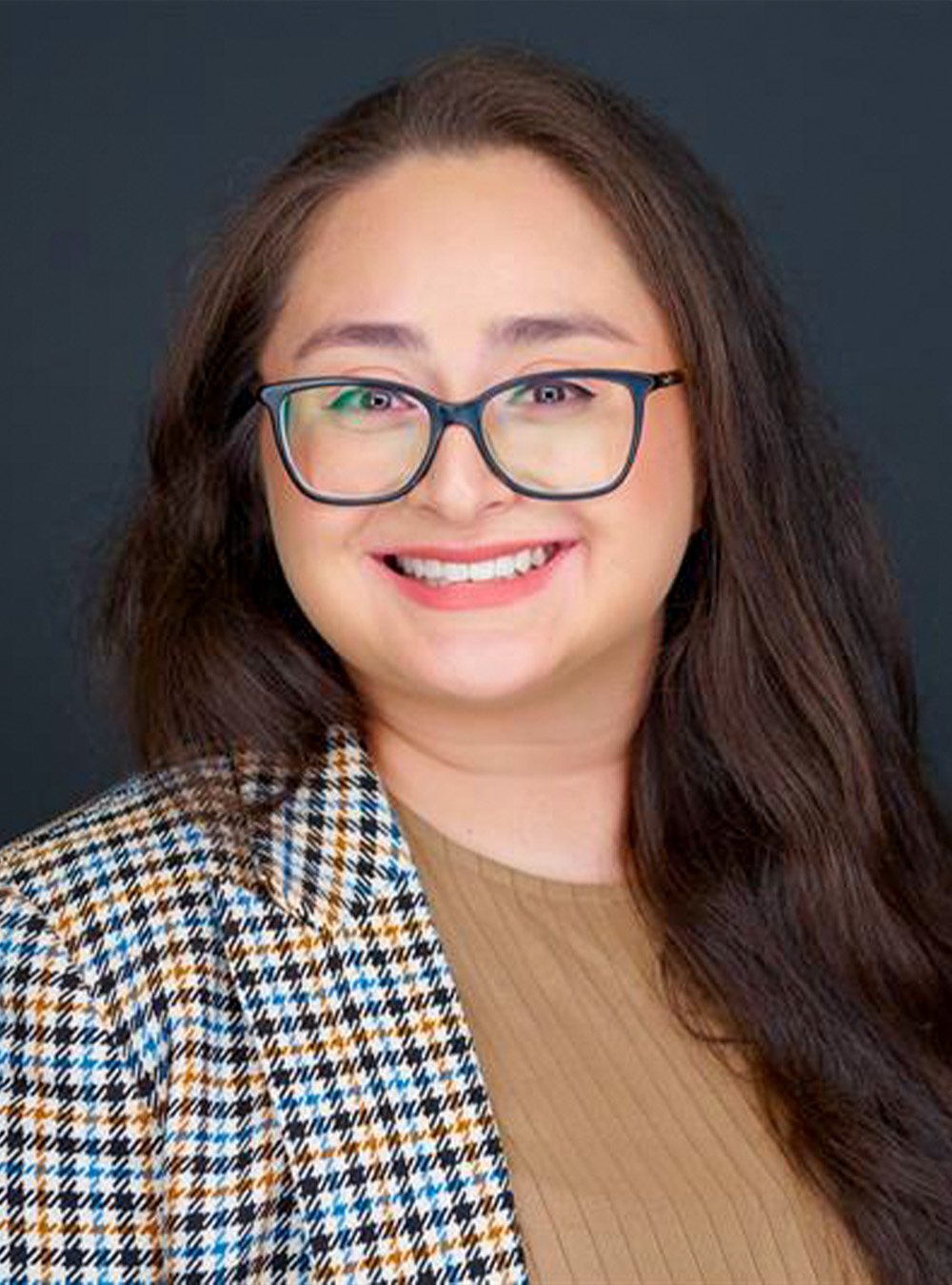 Portrait of a smiling woman with long dark hair and glasses wearing a patterned blazer against a dark background